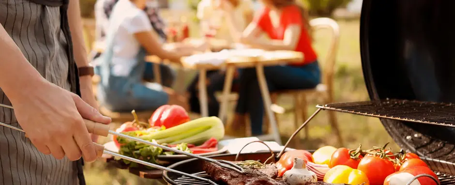 Man cooking steak on a grill, people sitting at a picnic table in the background
