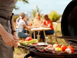 Man cooking steak on a grill, people sitting at a picnic table in the background